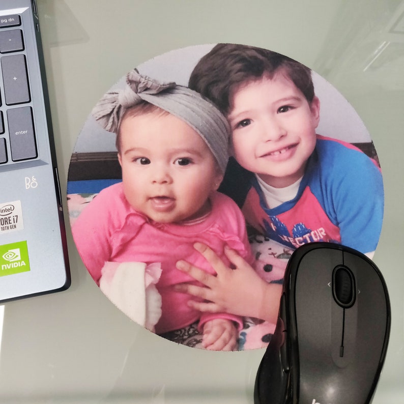 May include: Round mousepad featuring a color photograph of two children. The baby is wearing a pink shirt and gray headband. The older child is wearing a blue and red shirt. A black computer mouse is next to the mousepad.