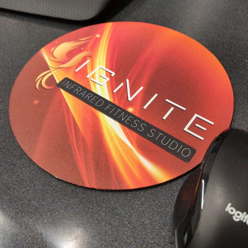 May include: A round mousepad with a fiery orange and red gradient design. The mousepad features the word "IGNITE" in white, along with the text "INFRARED FITNESS STUDIO". A black computer mouse is partially visible.