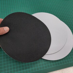 May include: A hand holds a black, circular sanding disc with a textured surface. Two additional gray sanding discs are visible. The discs are on a green cutting mat.