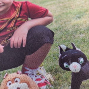 May include: A black cat plush toy with green eyes and white accents. The plush toy is on a green grassy background. A person is visible in the image, wearing a red shirt and black shorts.