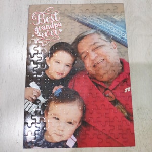 May include: A completed jigsaw puzzle featuring a family portrait. The puzzle shows a man in a red shirt with two children. The text "Best grandpa ever" is printed at the top. The puzzle is on a light-colored surface.