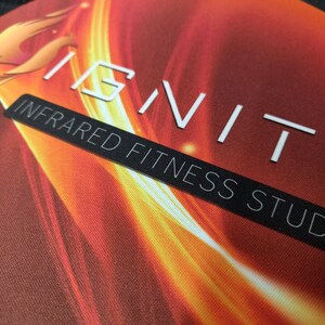 May include: Close-up of a fabric with the word "IGNIT" in white, above a black banner that reads "INFRARED FITNESS STUDIO". The background features a fiery orange and yellow design.