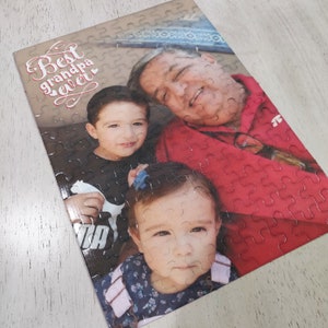May include: A completed jigsaw puzzle featuring a family portrait. The puzzle shows a man in a red shirt with two children. The text "Best grandpa ever" is printed in a decorative font on the top left corner.