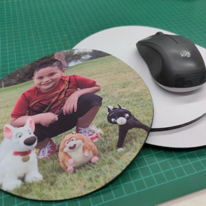 May include: Round mouse pad featuring a smiling child in a red shirt, with cartoon characters. A black wireless mouse sits on top of a white mouse pad. The background is a green cutting mat.