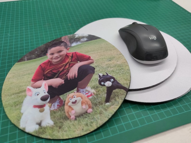 May include: Round mouse pad featuring a smiling child in a red shirt, with cartoon characters. A black wireless mouse sits on top of a white mouse pad. The background is a green cutting mat.