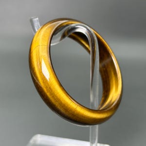 May include: A polished bangle bracelet crafted from golden-brown tiger's eye gemstone. The circular band features a smooth, reflective surface with natural chatoyant bands of light and dark amber tones that create a shimmering, silky appearance.