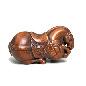 May include: A detailed wooden sculpture of a resting horse, featuring a saddle with intricate carvings. The horse is a rich brown color, with its head tucked down and legs folded beneath its body. The sculpture is set against a white background.