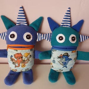 Può includere: Due bambole mostro di peluche con corpi blu e verde acqua, grandi occhi e corna a strisce. Uno ha una grafica di polpo, l'altro uno squalo pirata. Entrambi hanno cerniere arancioni e sono su una superficie bianca.