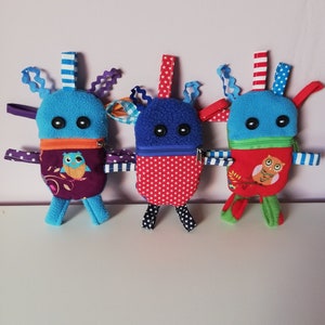 Könnte beinhalten: Drei bunte Plüschtiere in Monsterform mit verschiedenen Mustern und Farben. Jedes Spielzeug hat eine Reißverschlusstasche und ein kleines Eulenmotiv auf der Vorderseite.