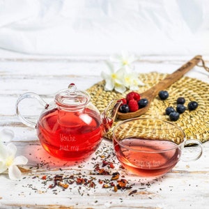 May include: A clear glass teapot and teacup filled with red tea. The teapot has the text "You're just my" etched on it, and the teacup says "cup of tea." Berries and a wooden spoon are also visible.