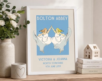 May include: A framed print featuring the text BOLTON ABBEY, VICTORIA & JOANNA, NORTH YORKSHIRE, 9TH JUNE 2019. The illustration depicts two figures in white dresses dancing against a light blue background, centred on a cream-coloured paper.