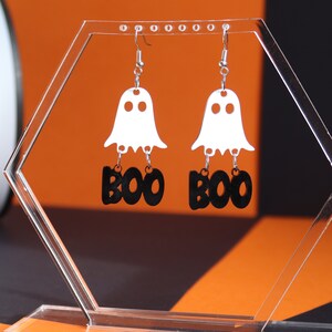May include: A pair of dangle earrings featuring white ghost silhouettes with black "BOO" lettering. The earrings are displayed on a clear acrylic stand.