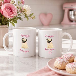May include: Two white ceramic mugs with the text "I puggin' love you" and "Puggin' love you too" in pink script. Each mug features a cartoon pug illustration. The mugs are on a marble surface with flowers and cookies.