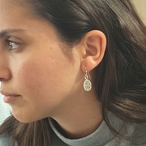 May include: A close-up of a person wearing silver dangle earrings with a delicate, intricate design. The earrings are small and feature a circular shape with a textured surface.