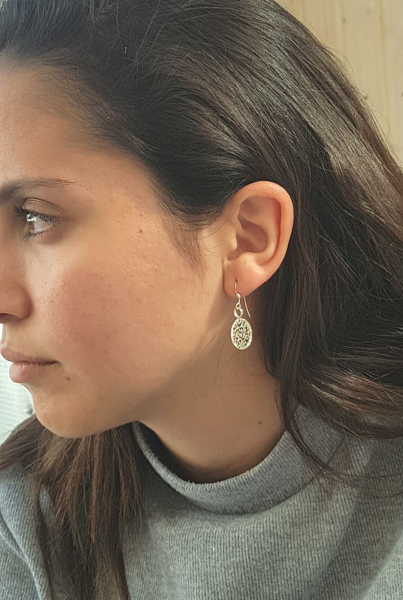May include: A close-up of a person wearing silver dangle earrings with a delicate, intricate design. The earrings are small and feature a circular shape with a textured surface.
