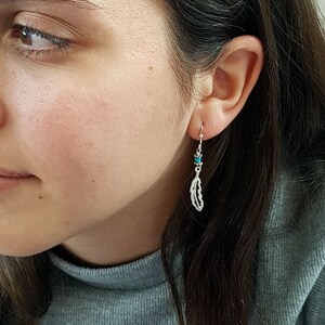 Op de afbeelding: Zilveren veren oorbellen met turquoise kralen. De oorbellen hangen aan de oorlel.
