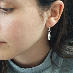 May include: A close-up of a person's ear with a silver dangle earring. The earring has a delicate, oval-shaped charm with a textured design.