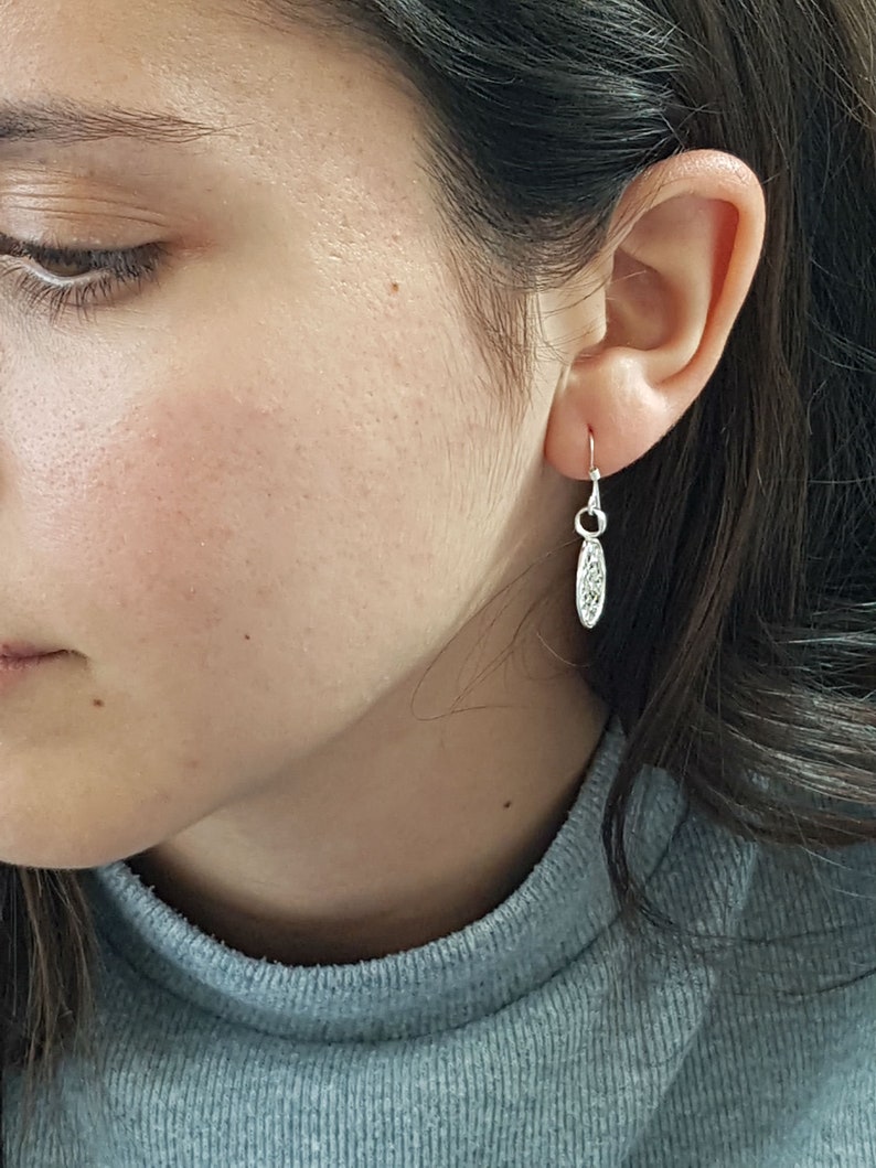 May include: A close-up of a person's ear with a silver dangle earring. The earring has a delicate, oval-shaped charm with a textured design.