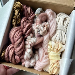 May include: A craft box filled with various shades of yarn and fabric scraps. The colors include dusty rose, cream, gold, and pale yellow. The box is open, revealing the assortment of textures and colors.