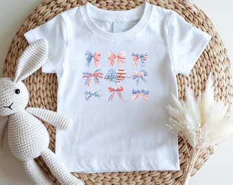 May include: White t-shirt featuring a design of patriotic bows in red, white, and blue. The bows are arranged in a grid pattern, with the text "4th July" in the center. The shirt is made of a soft, comfortable material.