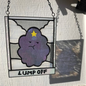 May include: Stained glass panel featuring a purple cloud character with a yellow star and a grumpy expression. The panel is framed in black and white, with the words "LUMP OFF" at the bottom. It hangs from a chain.