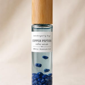 May include: A clear glass roller bottle with a light wood cap containing a pale blue liquid and small blue speckled stones. The white label reads: neurodivergent by design, COPPER PEPTIDE, roller serum, GHK-Cu, Hyaluronic Acid.