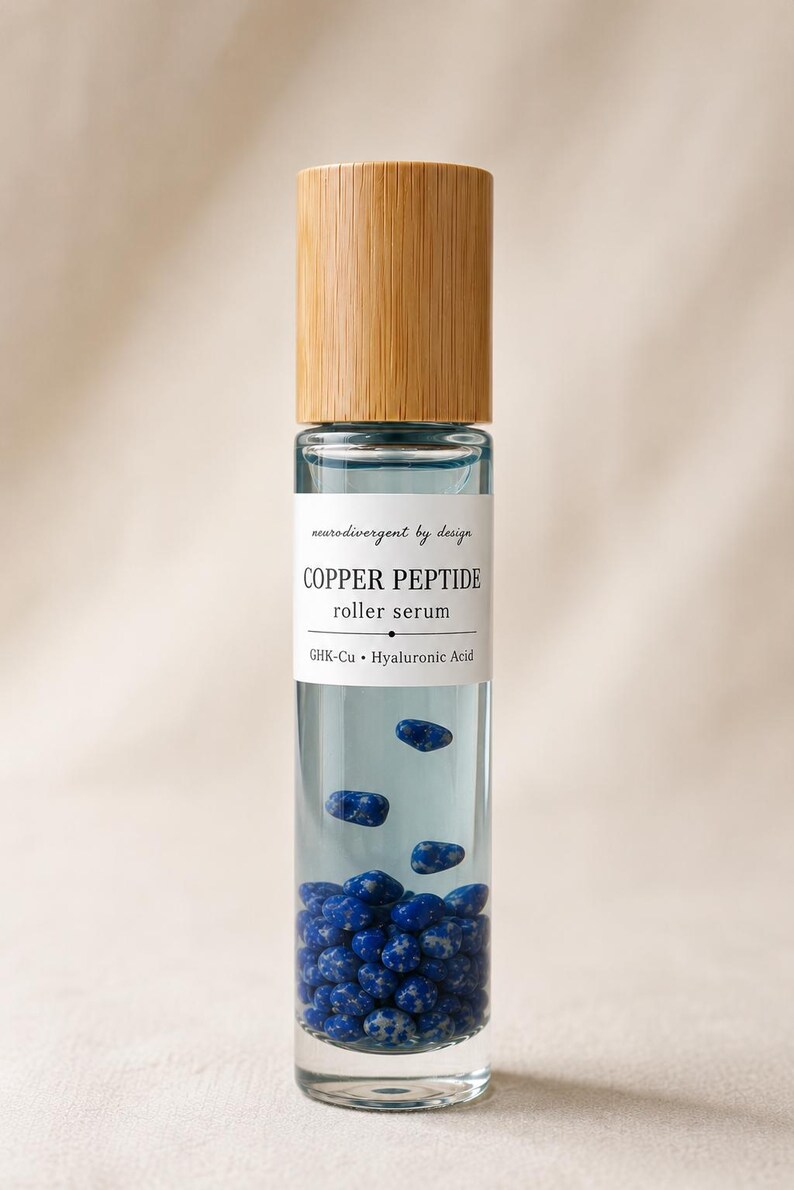May include: A clear glass roller bottle with a light wood cap containing a pale blue liquid and small blue speckled stones. The white label reads: neurodivergent by design, COPPER PEPTIDE, roller serum, GHK-Cu, Hyaluronic Acid.