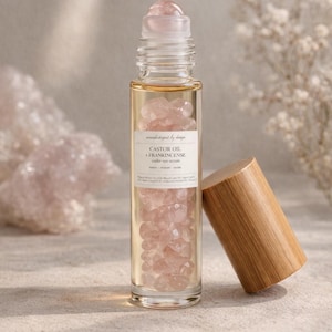 May include: A clear glass roller bottle filled with pink gemstones and a light-colored liquid. The bottle has a wooden cap resting beside it. The label reads "Castor Oil & Frankincense under eye serum."