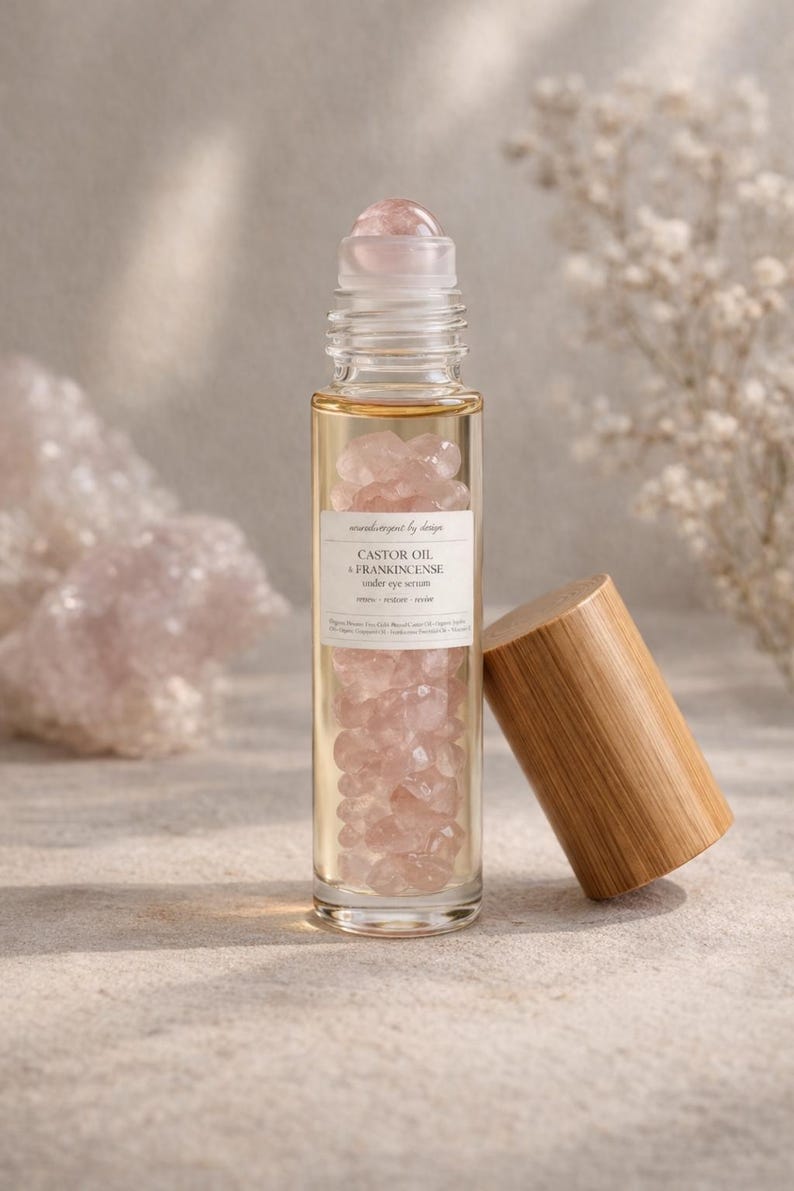 May include: A clear glass roller bottle filled with pink gemstones and a light-colored liquid. The bottle has a wooden cap resting beside it. The label reads "Castor Oil & Frankincense under eye serum."