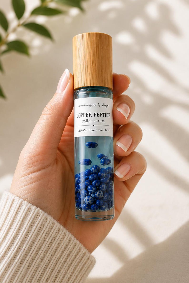 May include: A clear glass roller serum bottle with a light wood cap. The label reads neurodivergent by design COPPER PEPTIDE roller serum GHK-Cu Hyaluronic Acid. The bottle contains a clear liquid and numerous small, round, blue speckled beads.
