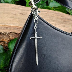 Könnte beinhalten: Ein silberner Schwertanhänger hängt an einem silbernen Verschluss, der an einer schwarzen Lederhandtasche befestigt ist. Der Schwertanhänger ist mit einem Parierstange und einer spitzen Klinge versehen. Die Handtasche hat eine geschwungene Form und einen schwarzen Riemen.