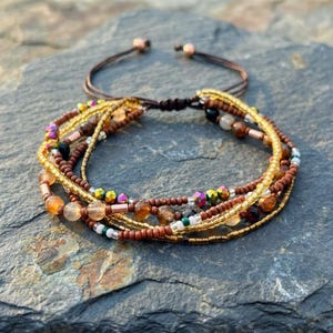 Puede incluir: Una pulsera de múltiples hilos con una mezcla de cuentas en tonos marrones, dorados e iridiscentes. La pulsera tiene un cierre de nudo corredizo ajustable. Las cuentas incluyen pequeñas cuentas de semillas y cuentas redondas más grandes.
