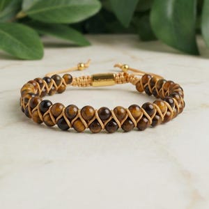 Könnte beinhalten: Ein zweireihiges Armband mit runden, polierten Tigeraugen-Perlen in Braun- und Goldtönen. Die Perlen sind mit einer beigefarbenen Kordel verflochten, und der verstellbare Verschluss enthält einen goldfarbenen Metallzylinder mit der Gravur by Santana.
