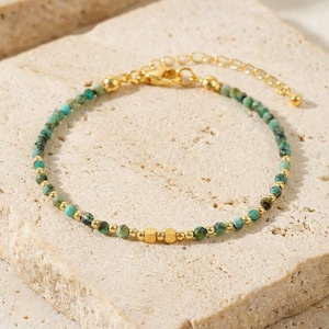 Könnte beinhalten: Zartes goldfarbenes Armband mit kleinen türkisfarbenen Perlen und goldenen Akzenten. Die verstellbare Kette und der Verschluss sind ebenfalls goldfarben. Das Armband wird auf einer neutralen Oberfläche präsentiert.