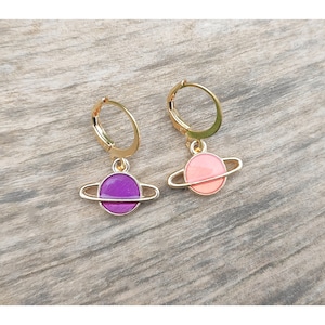 Könnte beinhalten: Ein Paar goldene Creolen mit einem kleinen Planeten-Anhänger. Ein Anhänger ist lila und der andere ist rosa.