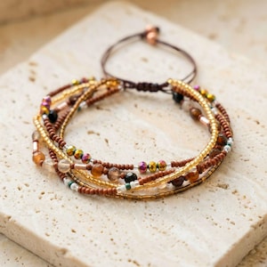 May include: A multi-strand beaded bracelet featuring a variety of earth-toned beads. The bracelet includes gold, brown, and iridescent beads, with a sliding knot closure. The beads are strung on a brown cord, creating a bohemian style.