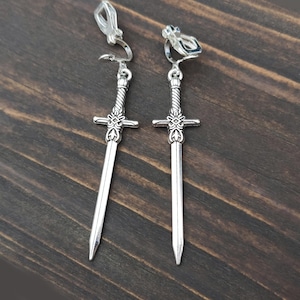 Pode incluir: Um par de brincos de pressão prateados em forma de espada. Cada brinco apresenta uma empunhadura detalhada com uma guarda cruzada e uma lâmina longa e pontiaguda. Os brincos são exibidos em uma superfície de madeira escura.