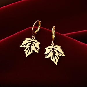 May include: A pair of gold-coloured leaf-shaped earrings. Each earring features a detailed leaf design suspended from a small hoop. The earrings are displayed against a deep red background, highlighting their metallic sheen.