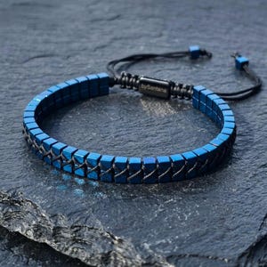 May include: A blue bracelet composed of small, square-shaped beads arranged in a single row. The beads are connected by black cord woven in a crisscross pattern. A black rectangular metal clasp features the engraved text bySantana.