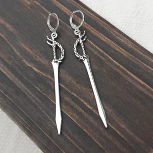 Pode incluir: Um par de brincos pendurados prateados. Cada brinco apresenta um design de espada com uma pega decorativa e uma lâmina longa e pontiaguda. Os brincos penduram-se de um fecho de argola simples. O fundo é uma superfície de madeira escura.