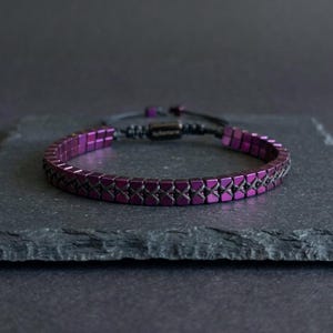 Op de afbeelding: Een armband met een enkele rij metallic paarse vierkante kralen in een herhalend patroon, met een zwart koord door het midden geweven. De armband heeft een verstelbare zwarte schuifsluiting met de merknaam Santoro erop gedrukt.