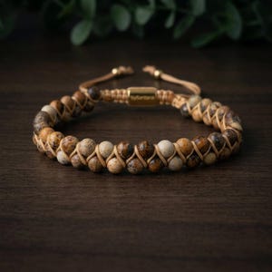 Op de afbeelding: Een armband met een mix van bruine, beige en crèmekleurige kralen. De kralen zijn geregen op een beige koord en de armband heeft een goudkleurige sluiting. De armband wordt gepresenteerd op een donker houten oppervlak.