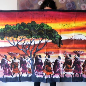 Puede incluir: Un tapiz de pared de batik colorido que representa a un grupo de personas que llevan cestas en la cabeza, caminando por un paisaje de sabana con un gran &aacute;rbol y una monta&ntilde;a en el fondo. El cielo es una puesta de sol naranja y amarilla vibrante.