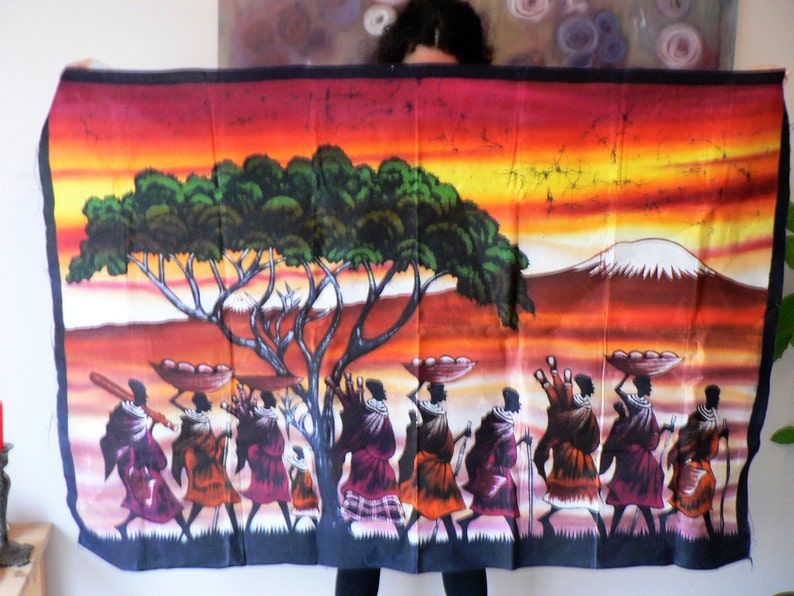 Puede incluir: Un tapiz de pared de batik colorido que representa a un grupo de personas que llevan cestas en la cabeza, caminando por un paisaje de sabana con un gran &aacute;rbol y una monta&ntilde;a en el fondo. El cielo es una puesta de sol naranja y amarilla vibrante.