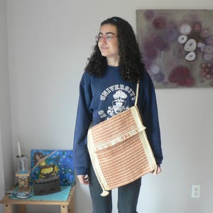 May include: A young person wearing a navy blue sweatshirt with the text "UNIVERSITY" on the front, and a brown and white woven bag slung over their shoulder.
