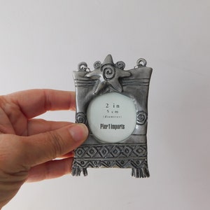 May include: A silver-toned metal picture frame with a starfish design and a 2-inch (5 cm) diameter opening. The frame has a decorative border and the text "Pier 1 Imports" is printed on the bottom.