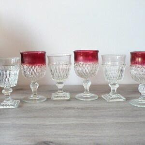 May include: Six clear glass goblets with a diamond pattern. Five of the goblets have a red rim. The goblets are arranged in a row on a wooden surface.