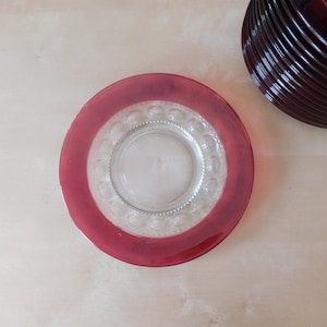 May include: A clear glass plate with a raised, textured rim and a red rim around the edge. The plate is empty.