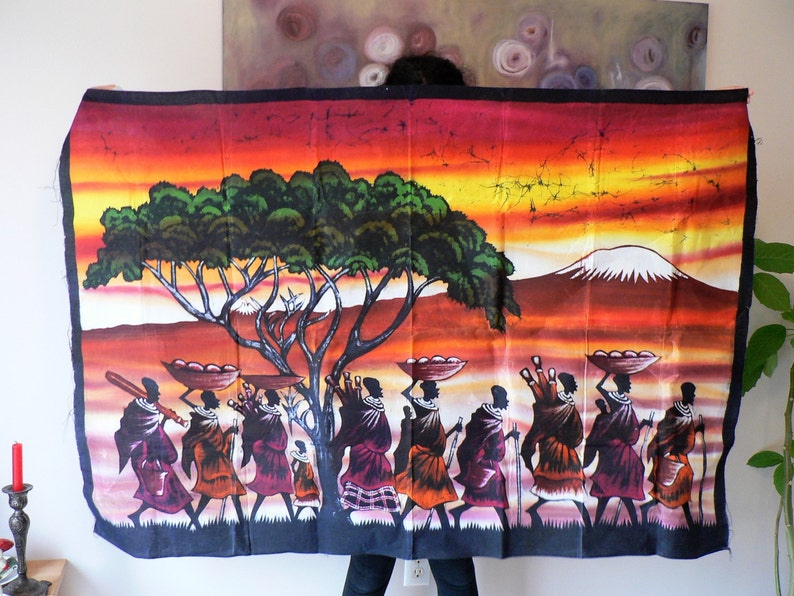 Puede incluir: Un tapiz de pared de batik colorido que representa a un grupo de personas que llevan cestas en la cabeza, caminando por un paisaje con un gran &aacute;rbol y una monta&ntilde;a en el fondo. La escena se desarrolla en un cielo de puesta de sol con tonos naranja, amarillo y rojo.