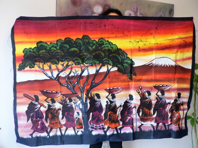 Puede incluir: Un tapiz de pared de batik colorido que representa una escena de personas caminando por un paisaje de sabana. La escena presenta un &aacute;rbol grande, una monta&ntilde;a en la distancia y un cielo de puesta de sol. Las personas llevan cestas y otros art&iacute;culos en sus cabezas.